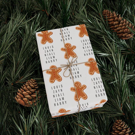 Gift wrapped in gingerbread-themed One Direction inspired wrapping paper featuring the names Louis, Zayn, Niall, Liam, and Harry in a repeating pattern, tied with twine and placed among pine branches and pinecones.