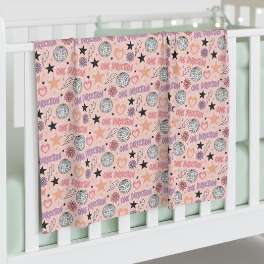 Pink One Direction inspired crib blanket draped over white crib rails featuring disco balls, stars, hearts, lightning bolts, and repeated “One Direction” pattern.
