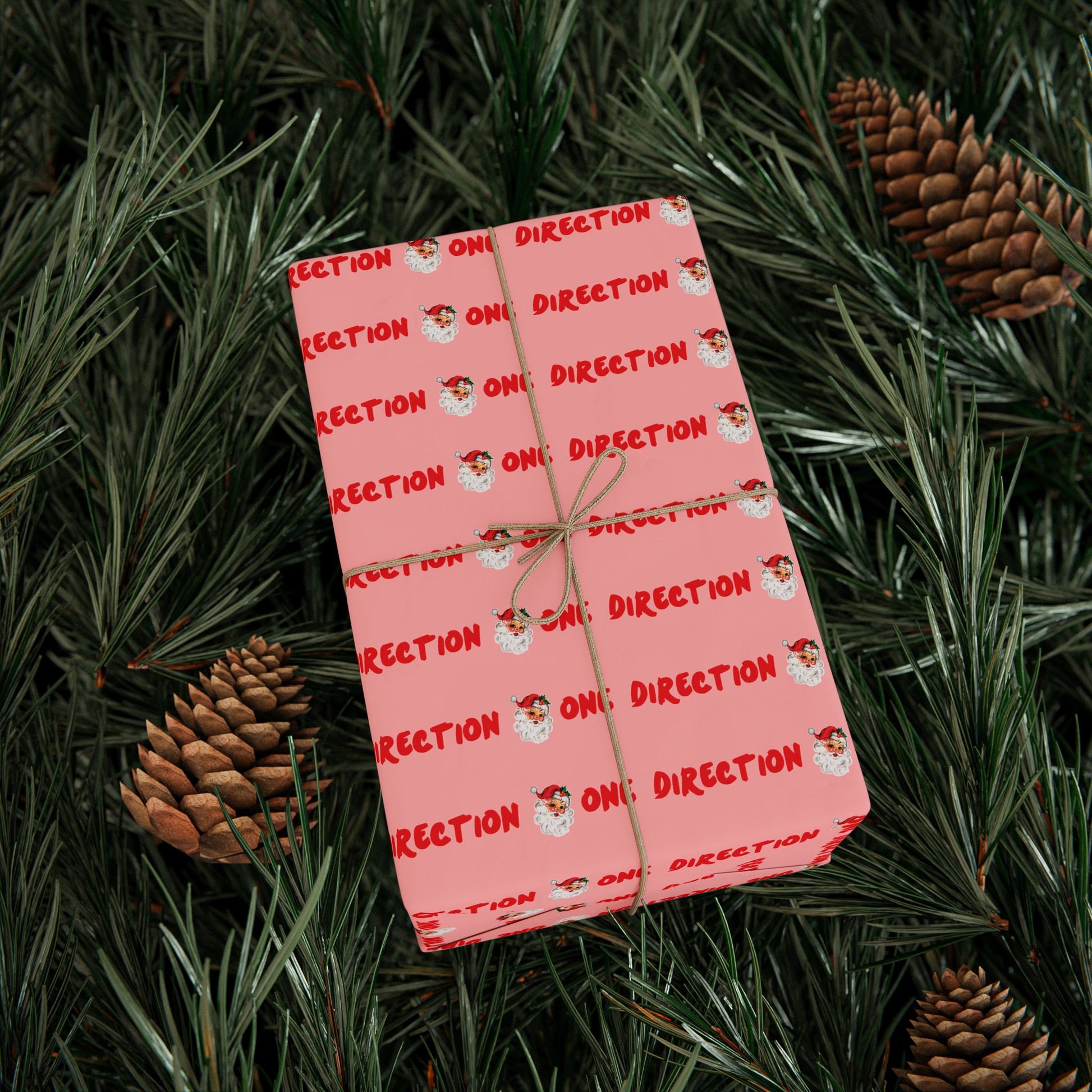 Pink fan-made One Direction inspired Christmas gift wrap featuring a repeating Santa Claus face pattern and red “One Direction” text, tied with twine and placed on pine branches with pinecones.