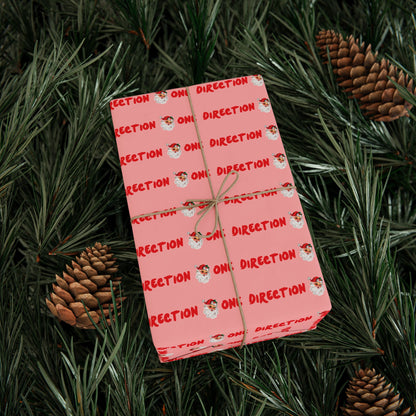 Pink fan-made One Direction inspired Christmas gift wrap featuring a repeating Santa Claus face pattern and red “One Direction” text, tied with twine and placed on pine branches with pinecones.