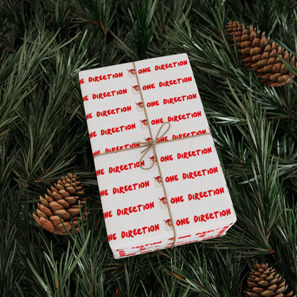 Pink fan-made One Direction inspired Christmas gift wrap featuring a repeating Santa Claus face pattern and red “One Direction” text, tied with twine and placed on pine branches with pinecones.