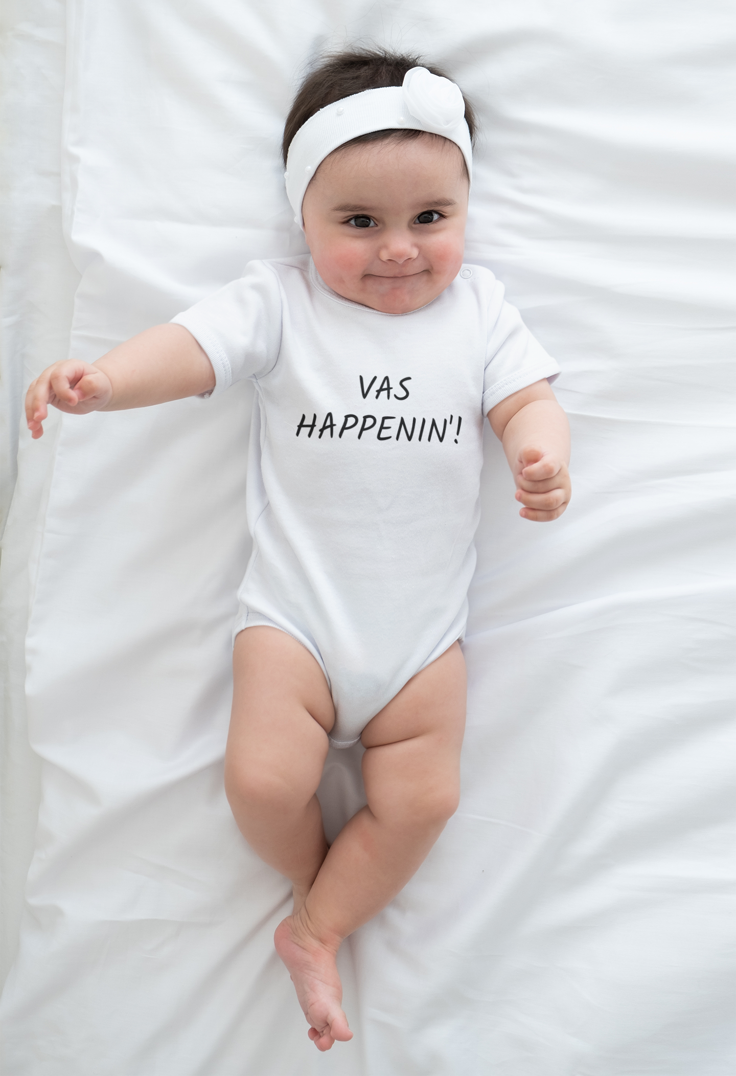 Baby wearing a white One Direction inspired onesie with the Zayn Malik quote “Vas Happenin’!” printed in black while lying on a white blanket.